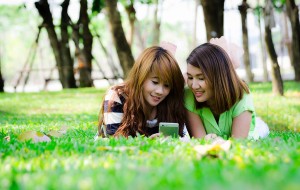 Two young Asian women lying down in the park looking at a mobile phone